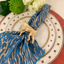 Load image into Gallery viewer, Decorative plate with a gold napkin holder shaped like a dog, blue and gold patterned napkin, and white flowers.