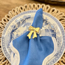 Load image into Gallery viewer, A golden bow napkin ring on a blue napkin, presented on a plate with a decorative border.
