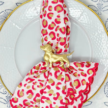 Load image into Gallery viewer, A napkin ring featuring a gold-colored metal wire shaped like a dachshund, placed on a napkin with a heart pattern, presented on a plate with a blue and white floral background.
